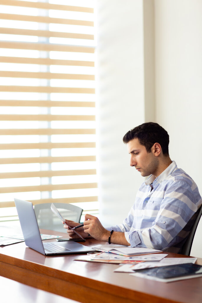 front view young handsome man striped shirt working inside conference hall using his silver laptop looking through documents writing down daytime work activity building